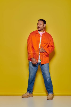 Photo for Full length of stylish indian man in orange jacket and denim jeans standing on yellow backdrop - Royalty Free Image