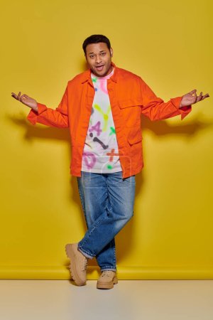 confident indian man in orange jacket and denim jeans standing with crossed legs on yellow backdrop