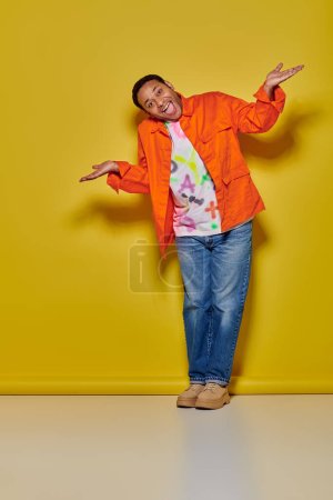 voller Länge positiver indischer Mann in orangefarbener Jacke und Jeans gestikulierend auf gelbem Hintergrund