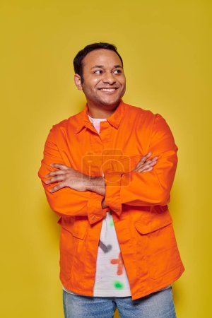 Foto de Retrato de hombre indio positivo en chaqueta naranja de pie con los brazos cruzados sobre fondo amarillo - Imagen libre de derechos