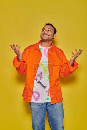 cheerful indian man in orange jacket and diy t-shirt gesturing on yellow backdrop, unbelievable