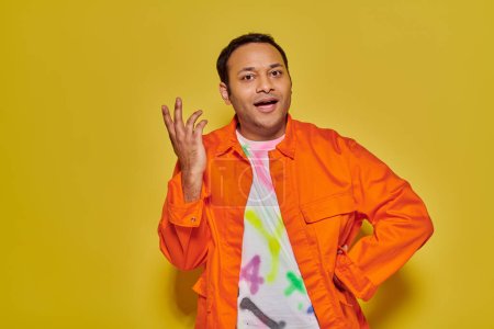 Foto de Hombre indio alegre en chaqueta naranja y camiseta bricolaje de pie con la mano en el lúpulo sobre fondo amarillo - Imagen libre de derechos