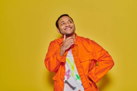 cheerful indian man in orange jacket and diy t-shirt standing with hand on hip on yellow backdrop