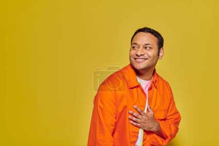 Porträt eines positiven indischen Mannes in orangefarbener Jacke, der wegschaut und auf gelbem Hintergrund lächelt