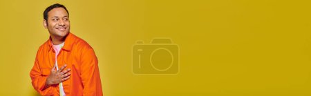 Téléchargez les photos : Portrait de l'homme indien positif en veste orange regardant loin et souriant sur fond jaune, bannière - en image libre de droit