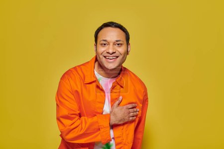 Foto de Retrato de hombre indio alegre en chaqueta naranja mirando a la cámara y sonriendo en el fondo amarillo - Imagen libre de derechos