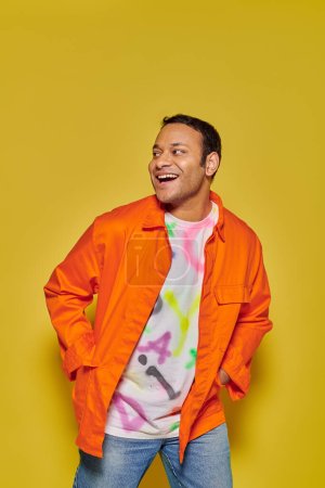 portrait of joyful indian man in orange jacket posing with hands in pockets on yellow backdrop