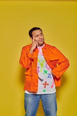 Photo for Portrait of pensive indian man in orange jacket posing with hand on hip on yellow background - Royalty Free Image