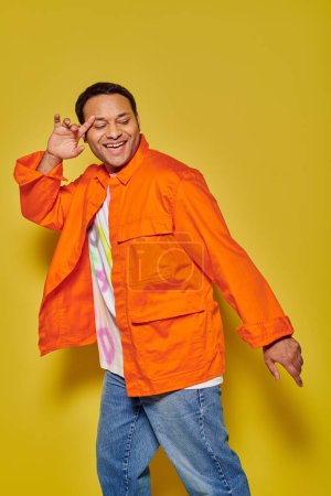 Porträt eines glücklichen indischen Mannes in orangefarbener Jacke und Jeansjacke, der auf gelbem Hintergrund tanzt