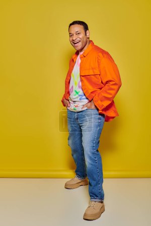 Foto de Longitud completa del hombre indio excitado en chaqueta naranja y chaqueta de mezclilla posando sobre fondo amarillo - Imagen libre de derechos