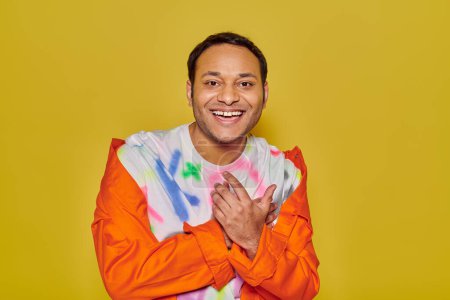 joyous indian man in orange jacket and diy t-shirt smiling and looking at camera on yellow backdrop
