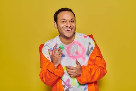 gleeful indian man in orange jacket and diy t-shirt smiling and looking at camera on yellow backdrop
