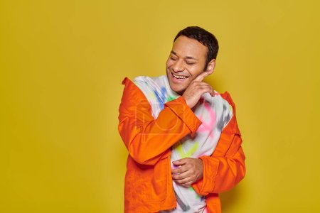 Photo for Gleeful indian man in orange jacket and diy t-shirt smiling with closed eyes on yellow backdrop - Royalty Free Image