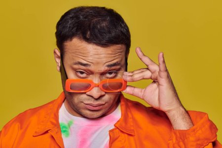 concepto de auto-expresión, hombre indio confiado ajustando gafas de sol naranjas sobre fondo amarillo