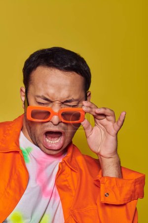 Foto de Expresión de la cara, hombre indio ajustando gafas de sol de color naranja y muecas en el fondo amarillo, actitud - Imagen libre de derechos