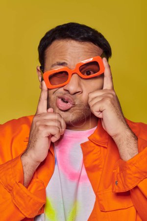 Foto de Expresión de la cara, hombre indio divertido ajustando gafas de sol de color naranja y muecas en el fondo amarillo - Imagen libre de derechos