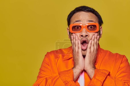 ekspresyjny, zszokowany Indianin w pomarańczowych okularach dotykający twarzy i mówiący wow na żółtym tle