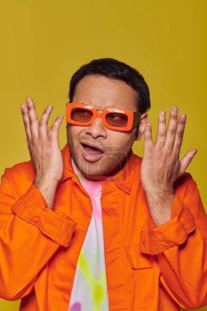 Verwirrter indischer Mann mit orangefarbener Sonnenbrille, wegschauend und gestikulierend auf gelbem Hintergrund, ausdrucksstark