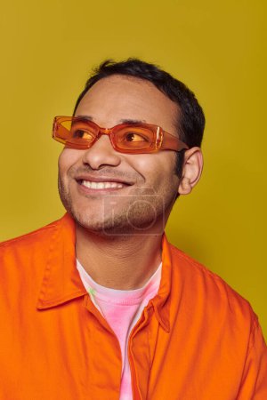 Foto de Hombre indio feliz en gafas de sol naranjas mirando hacia otro lado mientras sonríe sobre fondo amarillo, mirada lateral - Imagen libre de derechos