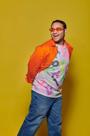 Foto de Hombre indio asombrado en gafas de sol y traje vibrante mirando hacia otro lado y sonriendo sobre fondo amarillo - Imagen libre de derechos