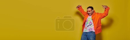 Téléchargez les photos : Homme indien en lunettes de soleil et tenue vibrante regardant vers le bas et gestuelle sur fond jaune, bannière - en image libre de droit