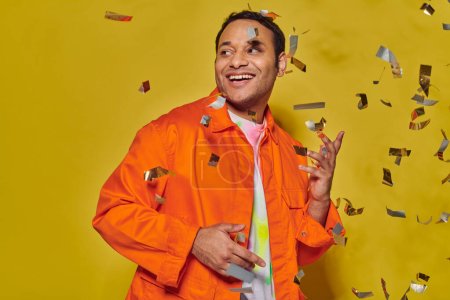 glücklicher indischer Mann in leuchtend orangefarbener Jacke lächelt neben fallendem Konfetti auf gelbem Hintergrund, Party