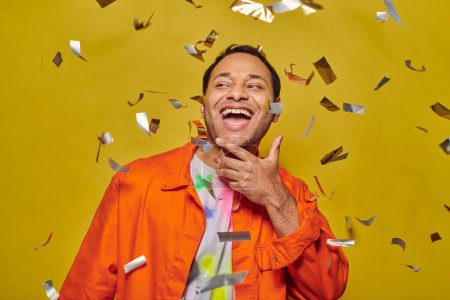 Photo for Joyous indian man in bright orange jacket smiling near falling confetti on yellow backdrop, party - Royalty Free Image