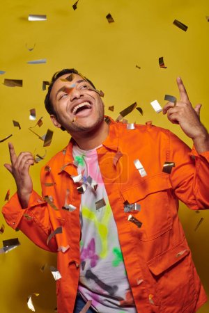 excited indian man in orange jacket gesturing near confetti on yellow backdrop, party concept