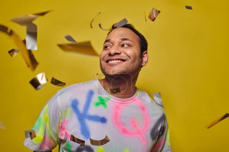 Foto de Complacido hombre indio en camiseta sonriendo cerca de caer confeti sobre fondo amarillo, concepto de fiesta - Imagen libre de derechos