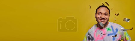 homme indien positif souriant près de tomber confettis sur fond jaune, concept de fête, bannière