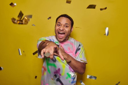 excited indian man pointing at camera near falling confetti on yellow backdrop, party concept