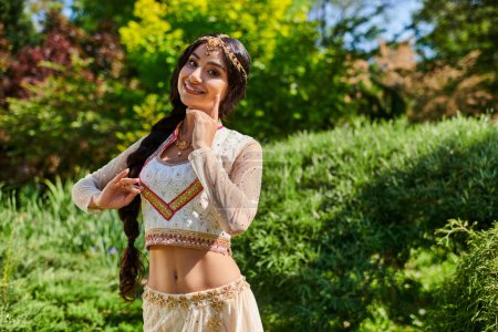 femme indienne gaie en tenue ethnique élégante posant à la caméra dans le parc d'été vert