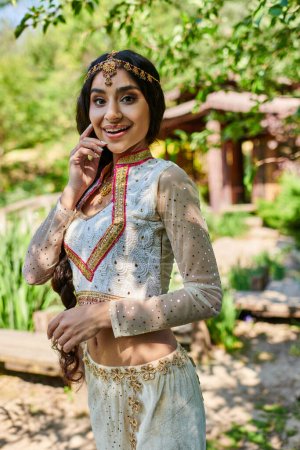 fröhliche asiatische Frau in lebendiger traditioneller Kleidung blickt im Sommerpark in die Kamera
