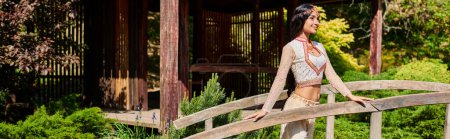 mujer india elegante joven en ropa auténtica de pie en el puente de madera en el parque de verano, pancarta