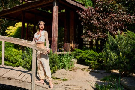 brünette indische Frau in lebendiger authentischer Kleidung schaut in der Nähe einer Holzbrücke im Sommerpark weg