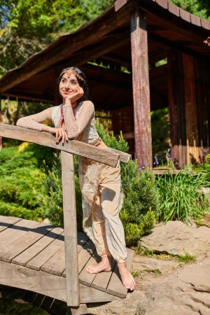mujer india feliz y soñadora en traje tradicional mirando hacia otro lado en el puente de madera en el parque de verano