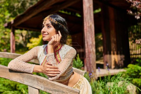 Foto de Parque de verano, soñadora mujer india de estilo auténtico sonriendo y mirando hacia otro lado en el puente de madera - Imagen libre de derechos