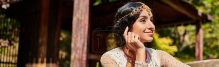 Photo for Brunette, smiling and dreamy indian woman in traditional clothes looking away in summer park - Royalty Free Image