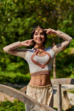 stylische indische Frau in traditioneller Kleidung posiert und lächelt an einem sonnigen Tag im Park in die Kamera