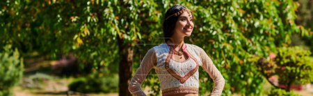 smiling indian woman in ethnic wear standing with hands on hips and looking away in park, banner