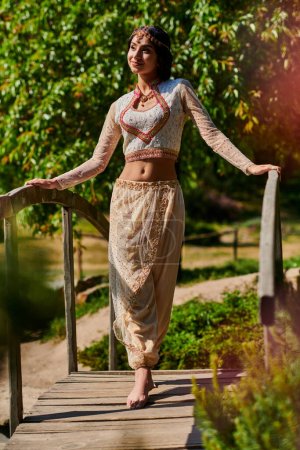 brunette indian woman in elegant traditional clothes standing on wooden bridge in sunny park