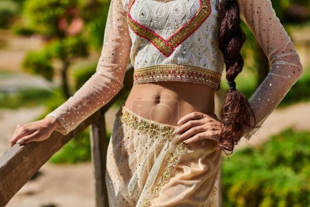 abgeschnittene Ansicht einer indischen Frau in traditioneller Kleidung, die mit der Hand auf der Hüfte im Sommerpark steht