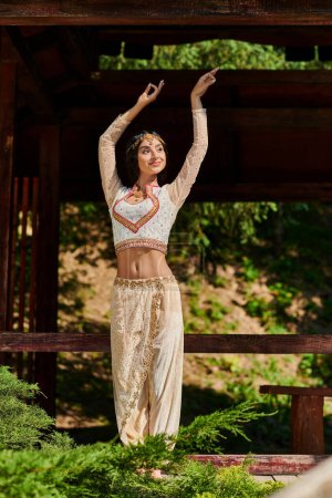fröhliche und anmutige indische Frau in authentischer Kleidung, die im Sommerpark tanzt und wegsieht