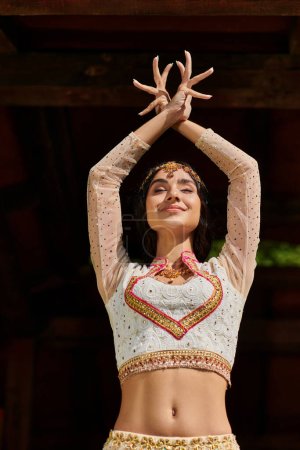 freudige junge Indianerin in eleganter ethnischer Kleidung tanzt mit geschlossenen Augen und erhobenen Händen im Freien