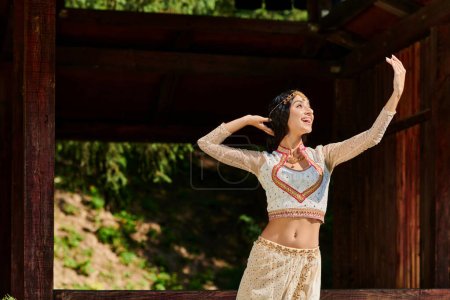Téléchargez les photos : Ravie et étonnée femme indienne en tenue traditionnelle regardant loin et dansant dans le parc en été - en image libre de droit