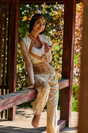 summer park, brunette indian woman in vibrant traditional clothes smiling at camera in wooden alcove