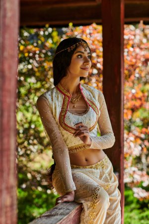 mujer india sonriente en traje de estilo auténtico sonriendo y mirando hacia otro lado en la alcoba de madera en el parque