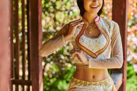 abgeschnittene Ansicht einer indischen Frau in eleganter und traditioneller Kleidung, die im Sommer im Park lächelt und tanzt