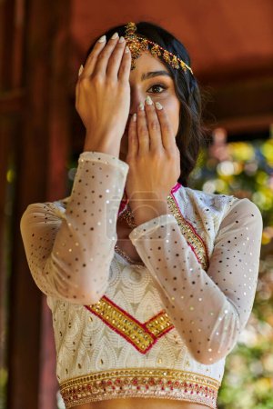 elegant indian woman in traditional clothes obscuring face with hands and looking at camera