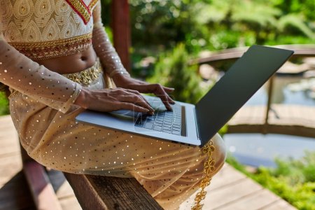 Photo for Indian woman in elegant traditional wear using laptop with blank screen on summer day, partial view - Royalty Free Image